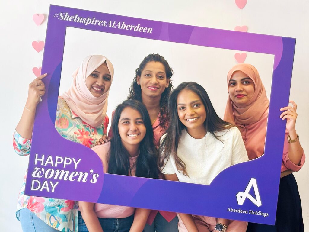 Alt text: A group of five smiling women from Aberdeen Holdings pose together holding a purple photo frame that reads “#SheInspiresAtAberdeen” and “Happy Women’s Day.” The frame also features the Aberdeen Holdings logo. The women are dressed in shades of pink and white, and pink heart decorations are visible in the background, celebrating International Women's Day.